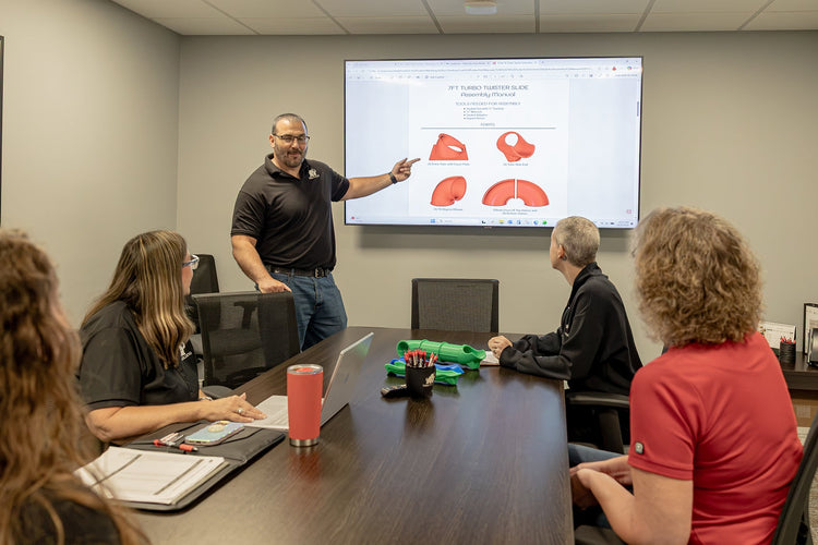 A man stands at the head of a conference room table, pointing to a large projection screen showing a diagram with red highlighted areas. Several people are seated around the table, and the room has gray walls, a dark wood conference table, and overhead fluorescent lighting.